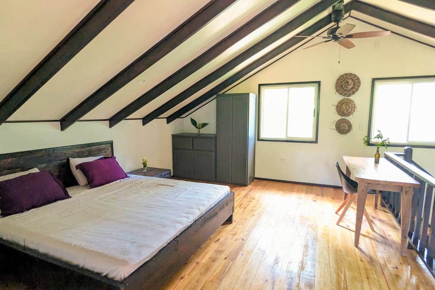 A bedroom with a bed, a desk, a chair, a ceiling fan, and a window.