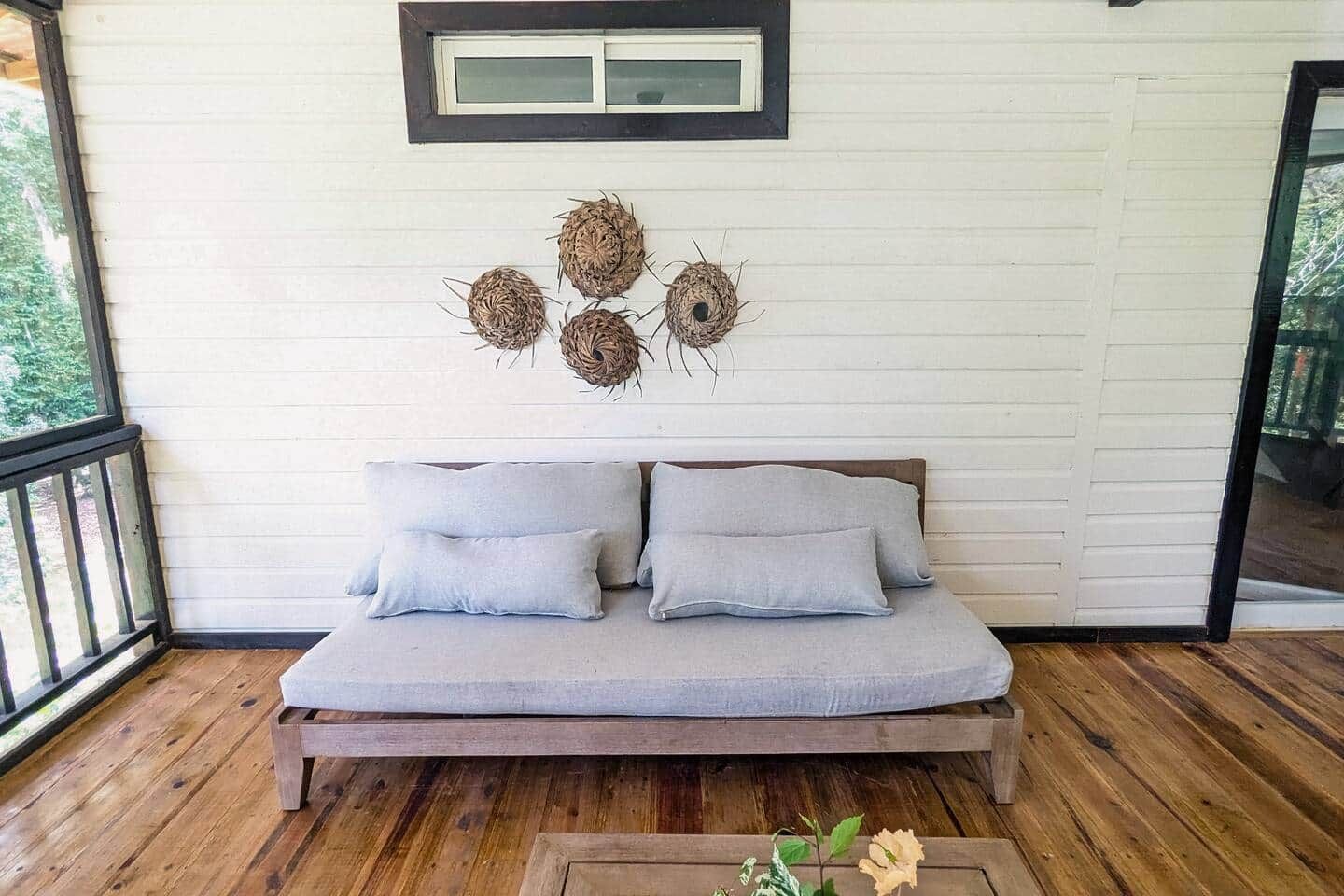 A wooden deck with a light grey sofa and cushions, a small table with flowers, and a wall with woven wall art.