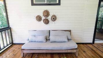 A wooden deck with a light grey sofa and cushions, a small table with flowers, and a wall with woven wall art.