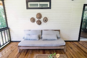 Eine Holzterrasse mit einem hellgrauen Sofa und Kissen, einem kleinen Tisch mit Blumen und einer Wand mit Wandbehängen.