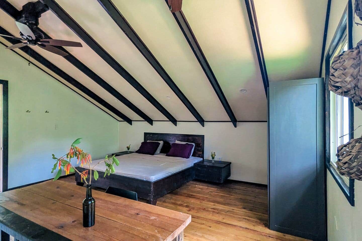 Un dormitorio con cama, mesita de noche, mesa de madera, ventilador de techo y ventana con cortinas.