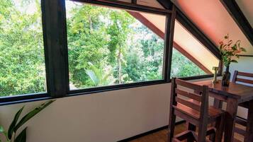 A room with a large window overlooking a dense forest, a wooden table, and a chair.