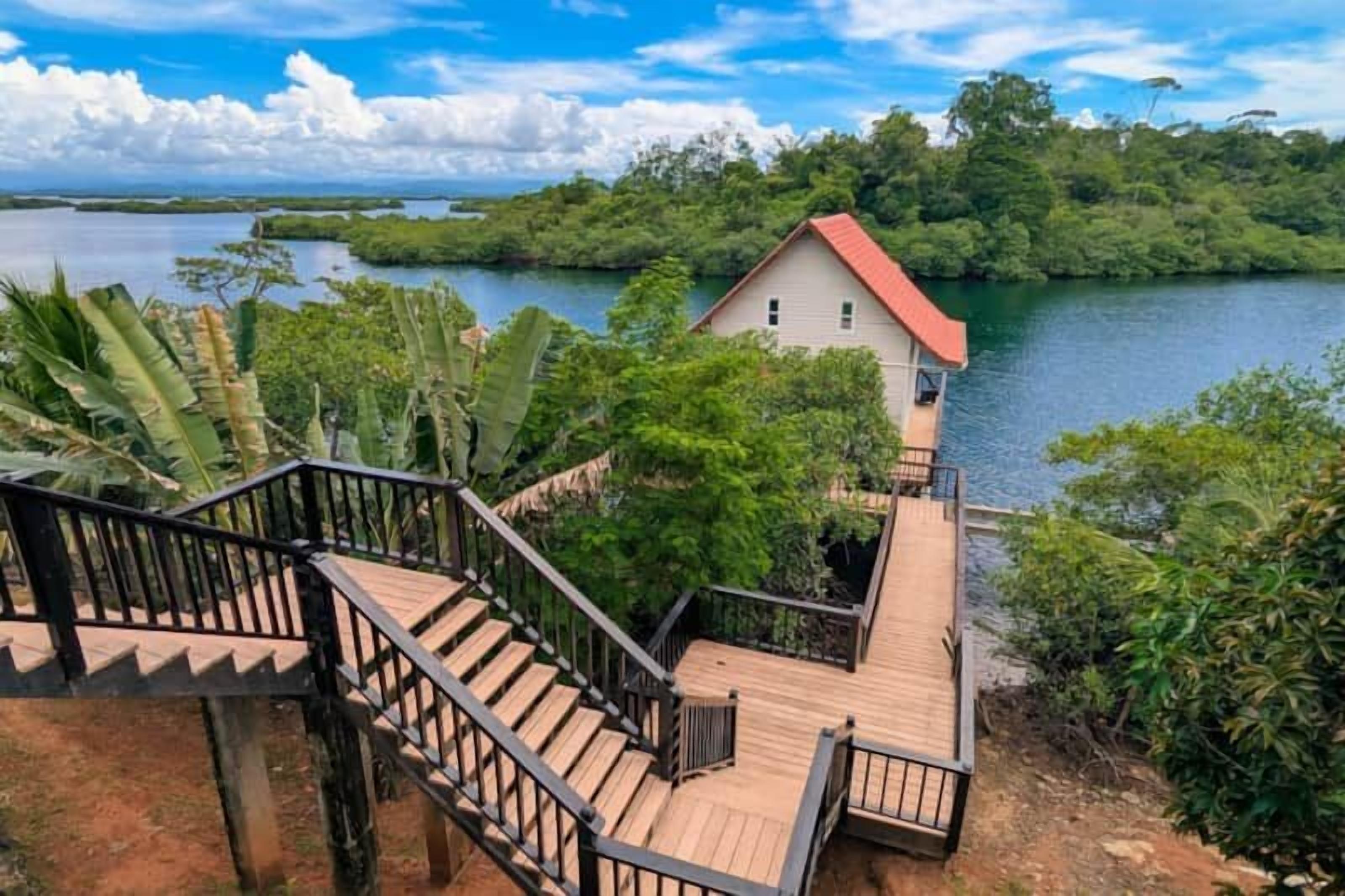 A wooden staircase leading to a house with a red roof, surrounded by lush greenery and a body of water.