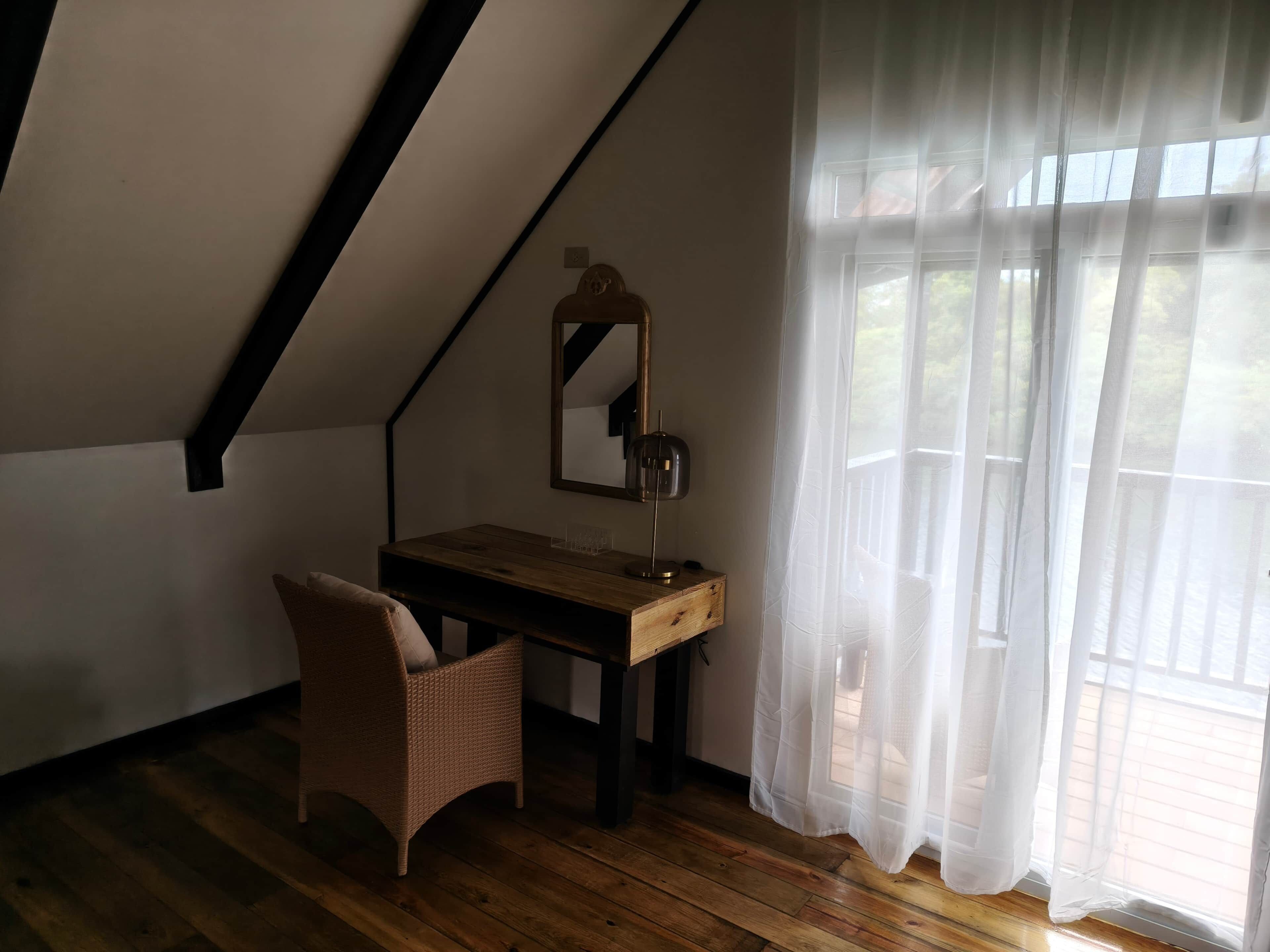 Una habitación con techo inclinado, un escritorio de madera, una silla, un espejo y una ventana con cortinas traslúcidas.