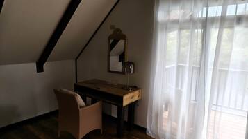 Una habitación con techo inclinado, un escritorio de madera, una silla, un espejo y una ventana con cortinas traslúcidas.
