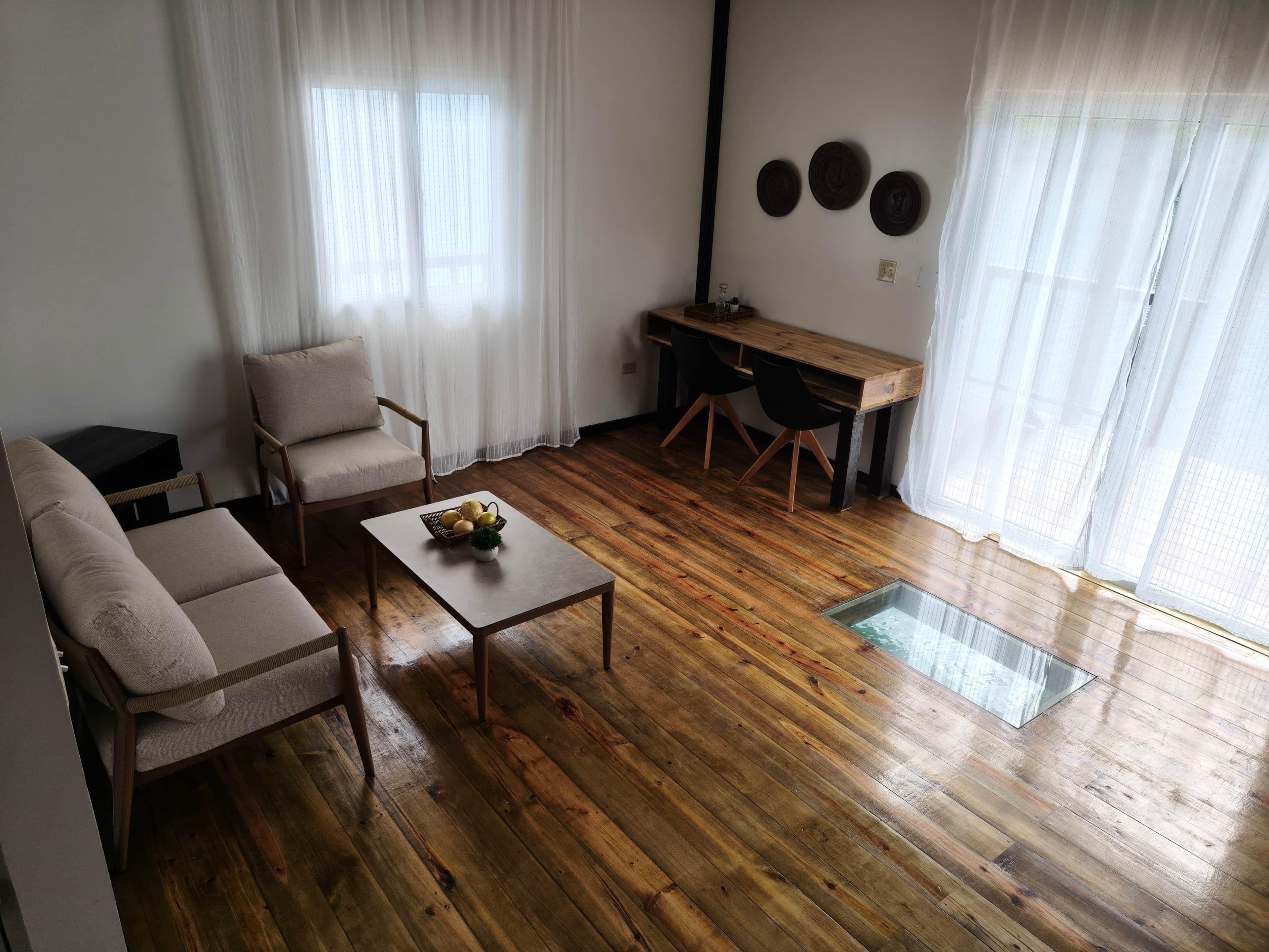 A living room with a sofa, a coffee table, a desk, and a chair. There are three wall-mounted decorations and a window with sheer curtains.