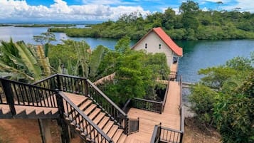 A wooden staircase leading to a house with a red roof, surrounded by lush greenery and a body of water.