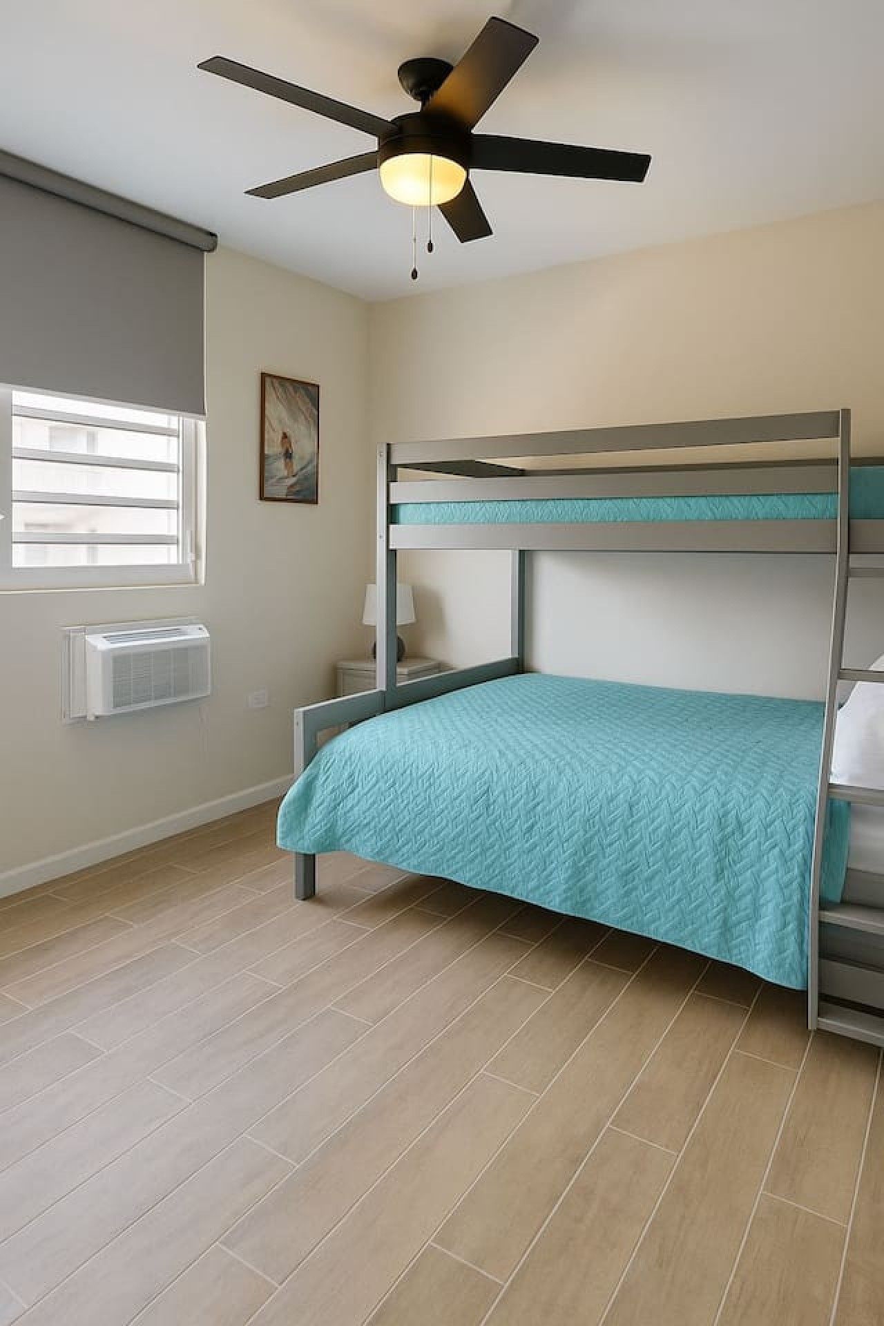 Una cama litera con colcha color turquesa, un ventilador de techo, una ventana con persianas y un aire acondicionado instalado en la pared.