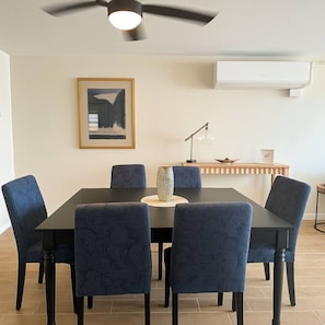A dining area with a black table, six blue upholstered chairs, a ceiling fan, a framed picture on the wall, and a small side table with a lamp and decorative items. - Costa Córcega 5A | Beachfront, Pool & Beach Access (Rincón)