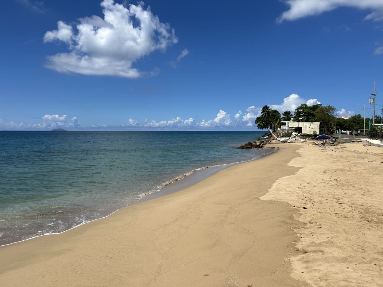 A beach with clear skies, calm water, and a few buildings in the background.