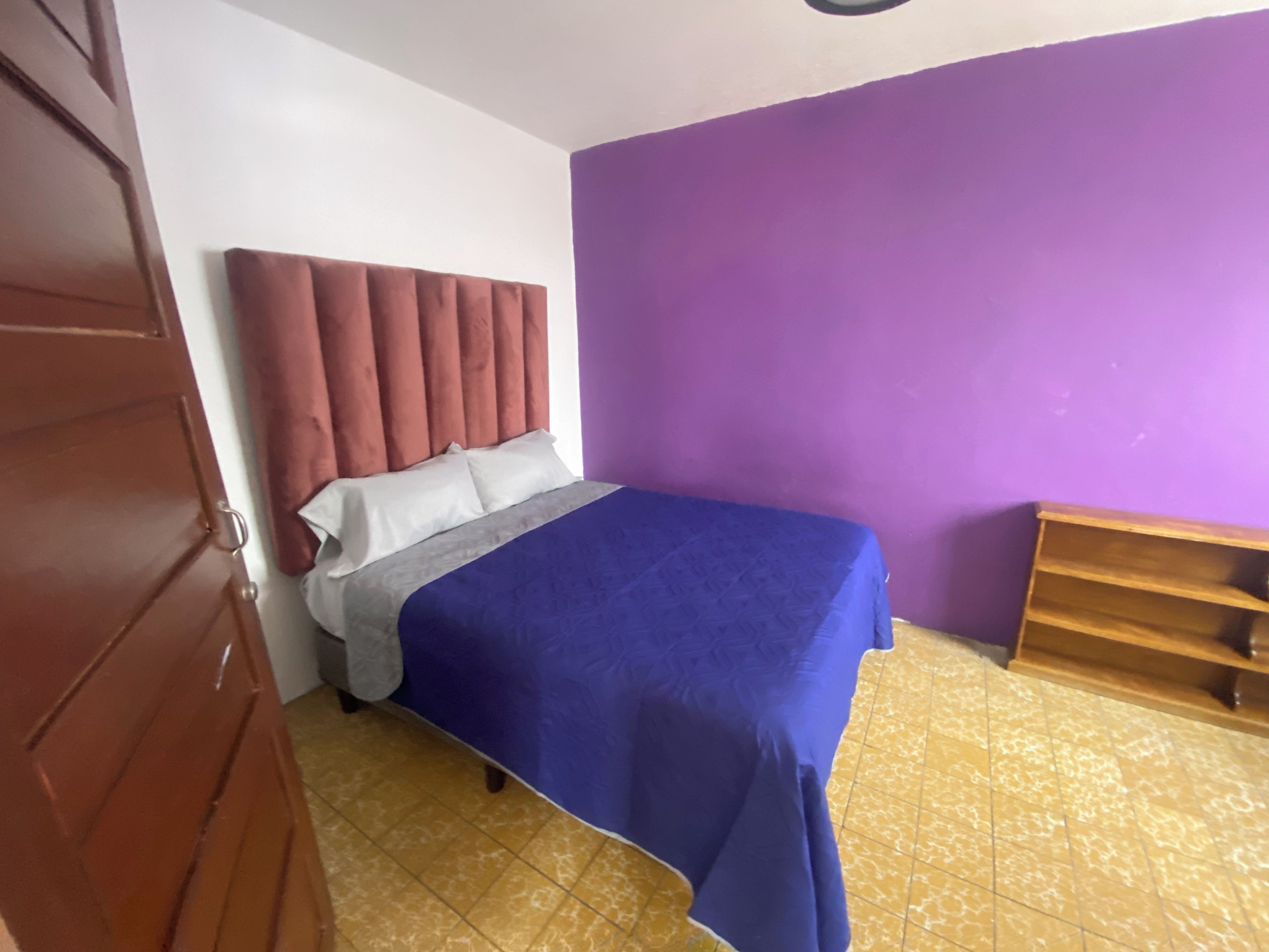 A bedroom with a bed, a headboard, a purple wall, a wooden shelf, and a tiled floor.
