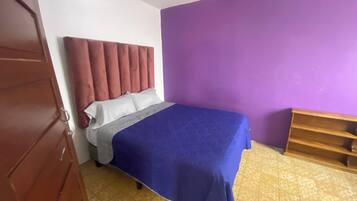 A bedroom with a bed, a headboard, a purple wall, a wooden shelf, and a tiled floor.
