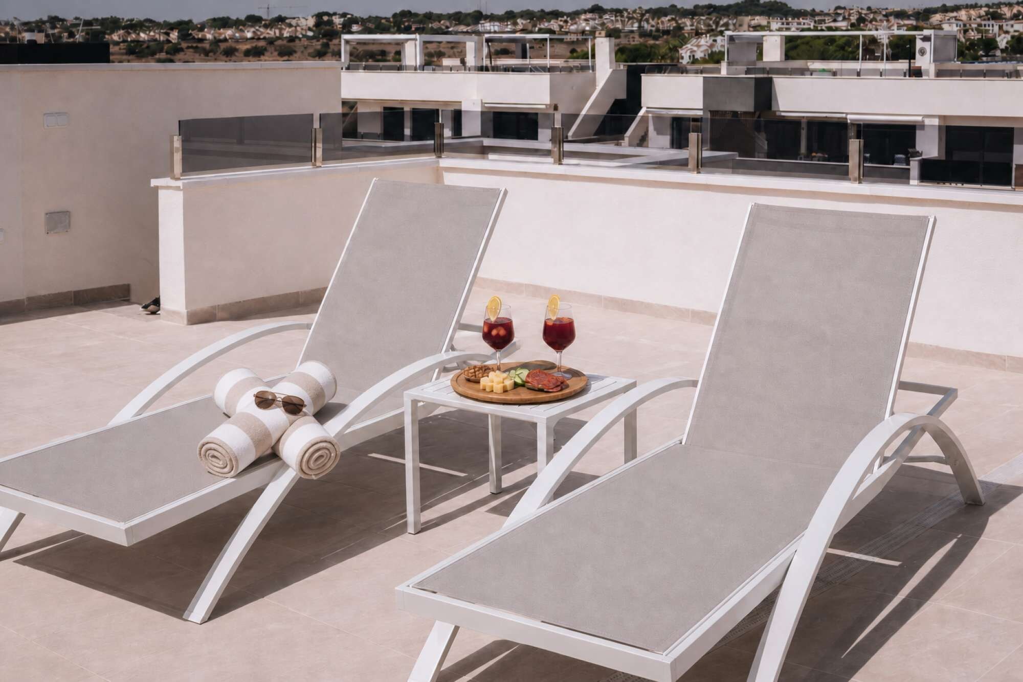 A rooftop terrace with two lounge chairs, a small table with a plate of food and drinks, and a view of the cityscape in the background.