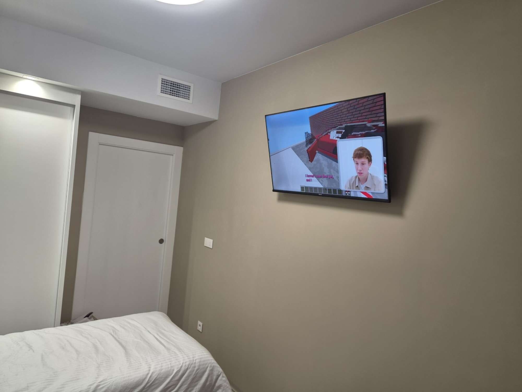 Een slaapkamer met een bed, een aan de muur bevestigde televisie en een deur.