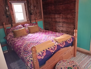 Un lit en bois avec une couvre-lit à motifs et deux oreillers, un petit tapis rond et une fenêtre donnant sur l’extérieur.