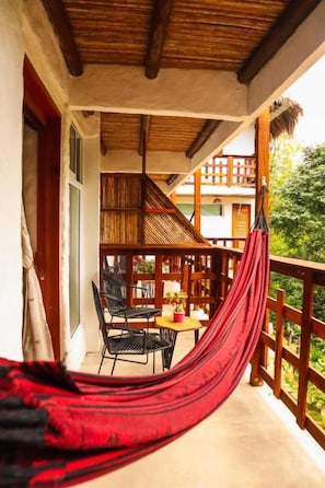 Un hamac sur un balcon doté d’une balustrade en bois, une table avec des chaises et une vue sur un bâtiment au toit de chaume.