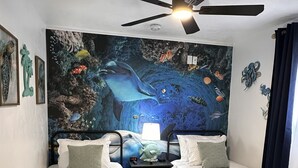 Una stanza con due letti singoli, un ventilatore a soffitto, un affresco raffigurante una scena subacquea e un comodino con una lampada.