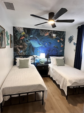 Una habitación con dos camas individuales, un ventilador de techo, un mural en la pared que representa una escena submarina y una mesita de noche con lámpara.