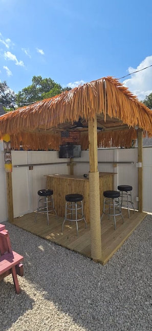 Zona de bar al aire libre con techo de paja, cuatro taburetes y una pantalla de televisión.