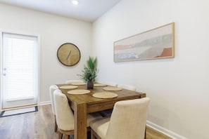 A dining area with a wooden table, six chairs, a clock, a potted plant, and a framed picture on the wall.