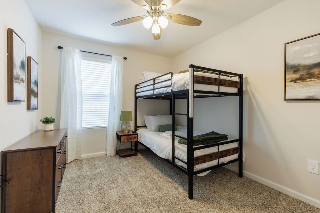 A bunk bed room with a ceiling fan, a bedside table with a lamp, and a window with curtains.