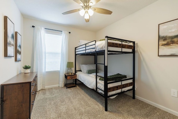 A bunk bed room with a ceiling fan, a bedside table with a lamp, and a window with curtains.