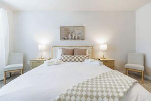 Quarto de hotel com uma cama grande, duas luminárias de cabeceira, um quadro na parede e duas cadeiras.