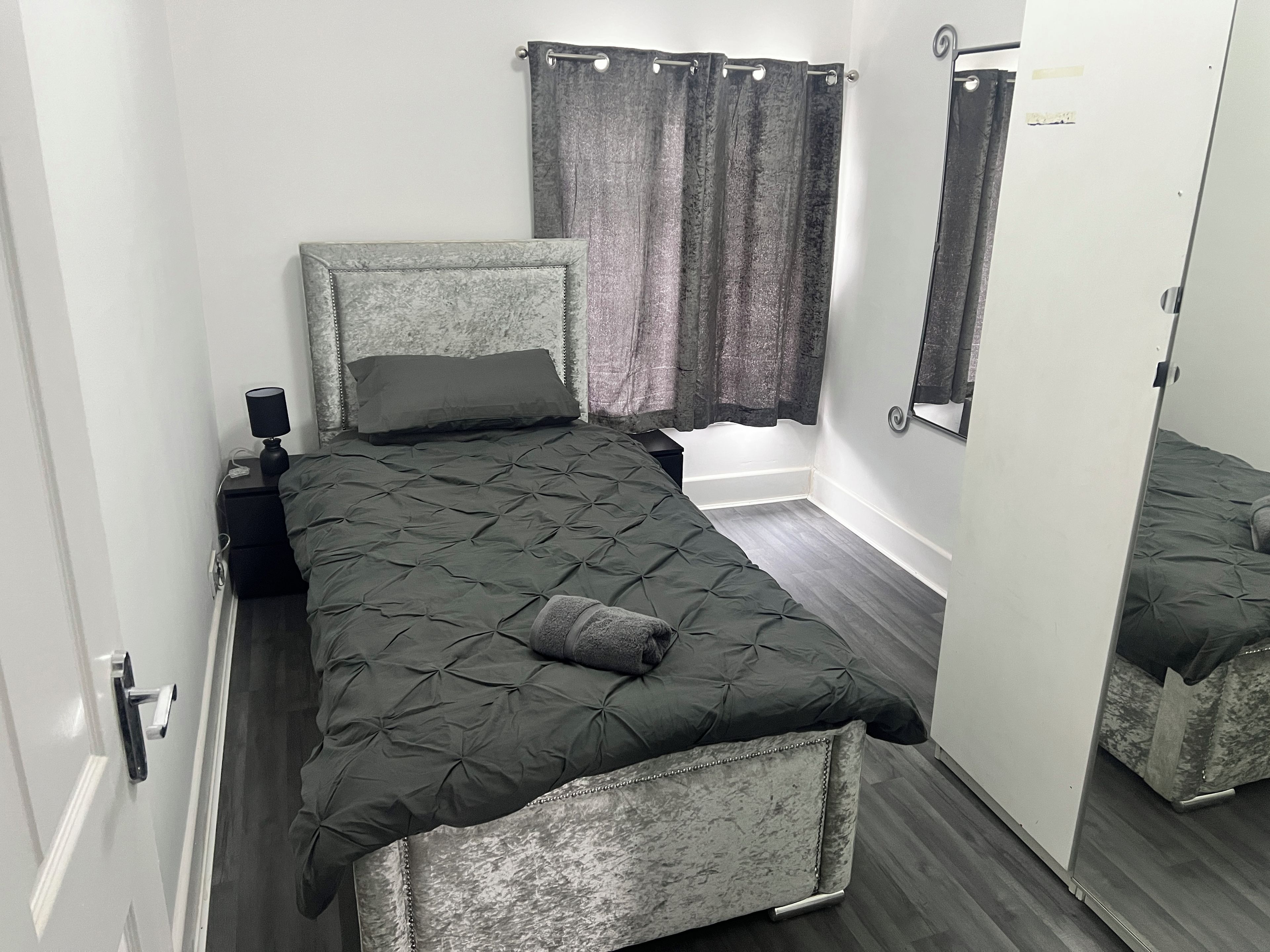 A bedroom with a bed, nightstand, lamp, curtains, and a mirror.