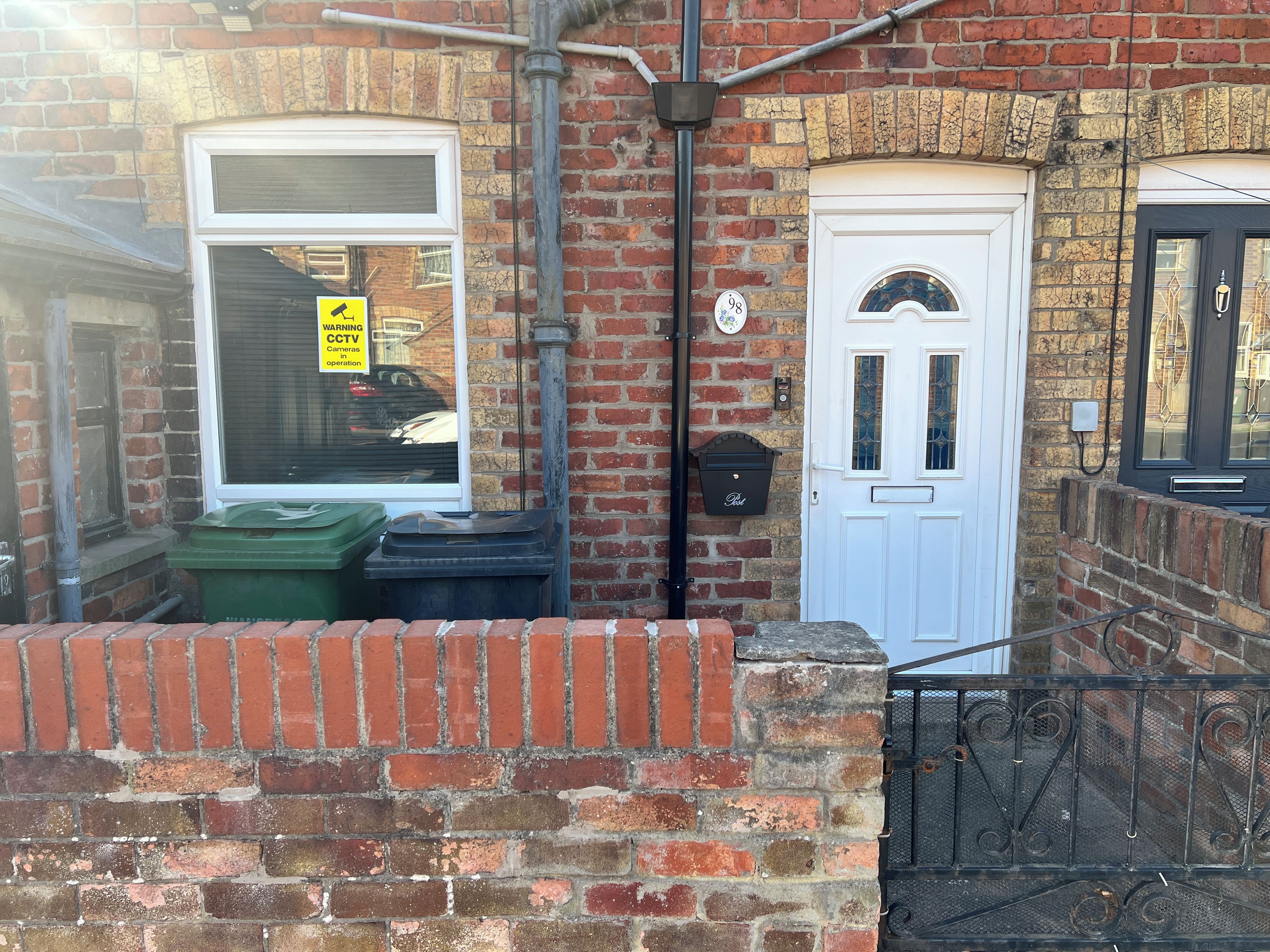 A brick building with a white door, a window with a warning sign, and a green bin outside.