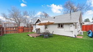 Patio trasero residencial con césped, una parrilla, una mesa de patio con sillas y una valla de madera.
