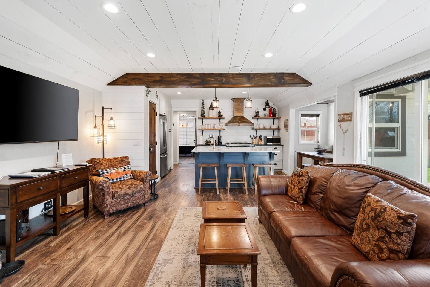 A spacious living room with a brown leather sofa, a patterned armchair, and a wooden coffee table. A flat-screen TV is mounted on the wall, and the room features a high ceiling with exposed wooden beams.