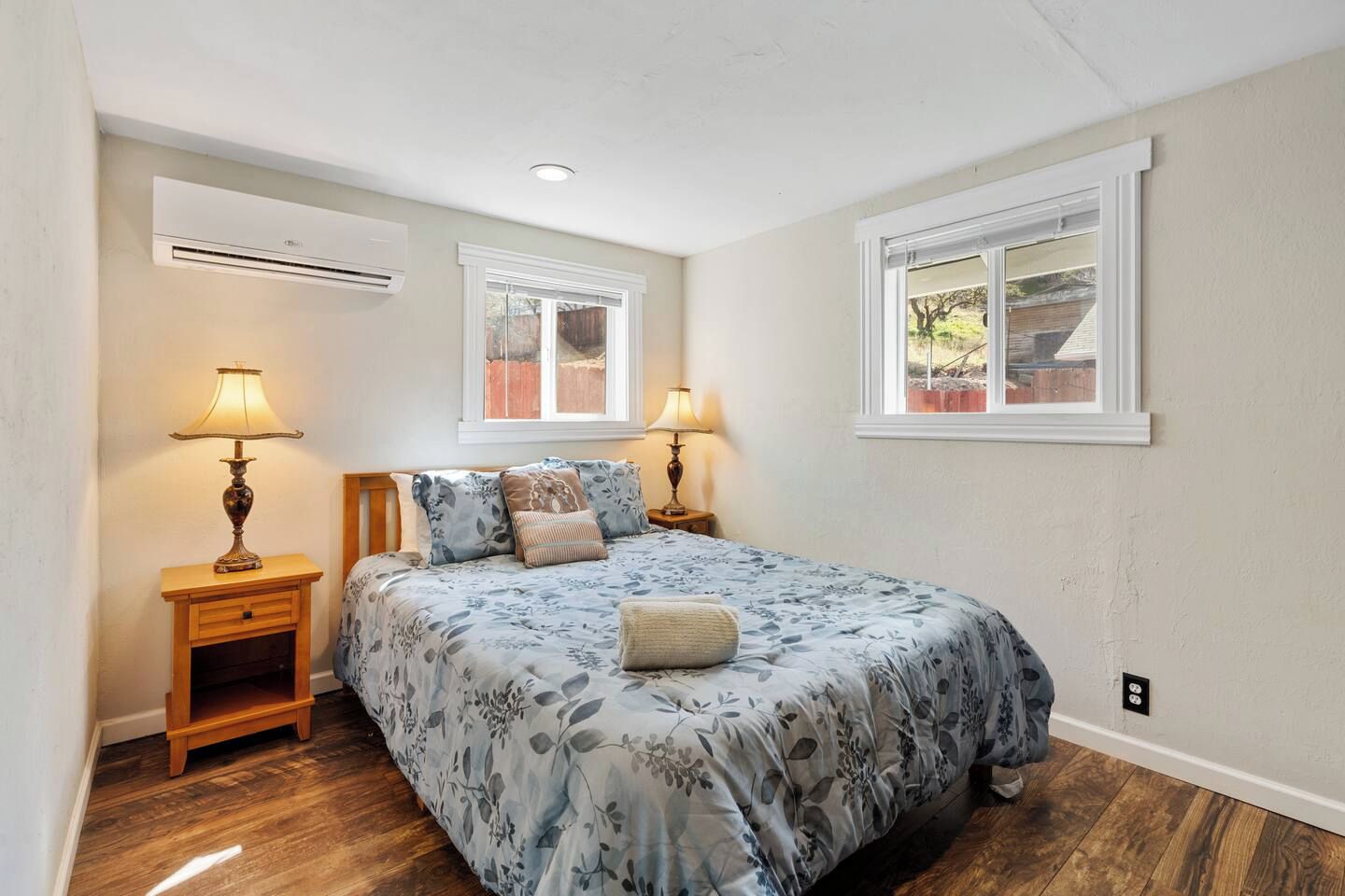 A bedroom with a bed, two bedside lamps, an air conditioning unit, and two windows.