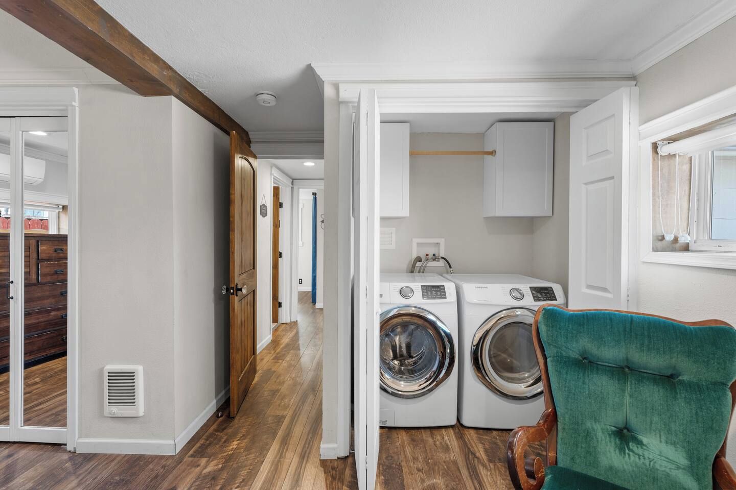 A laundry room with a wooden floor, two washing machines, a green chair, and a window with curtains.
