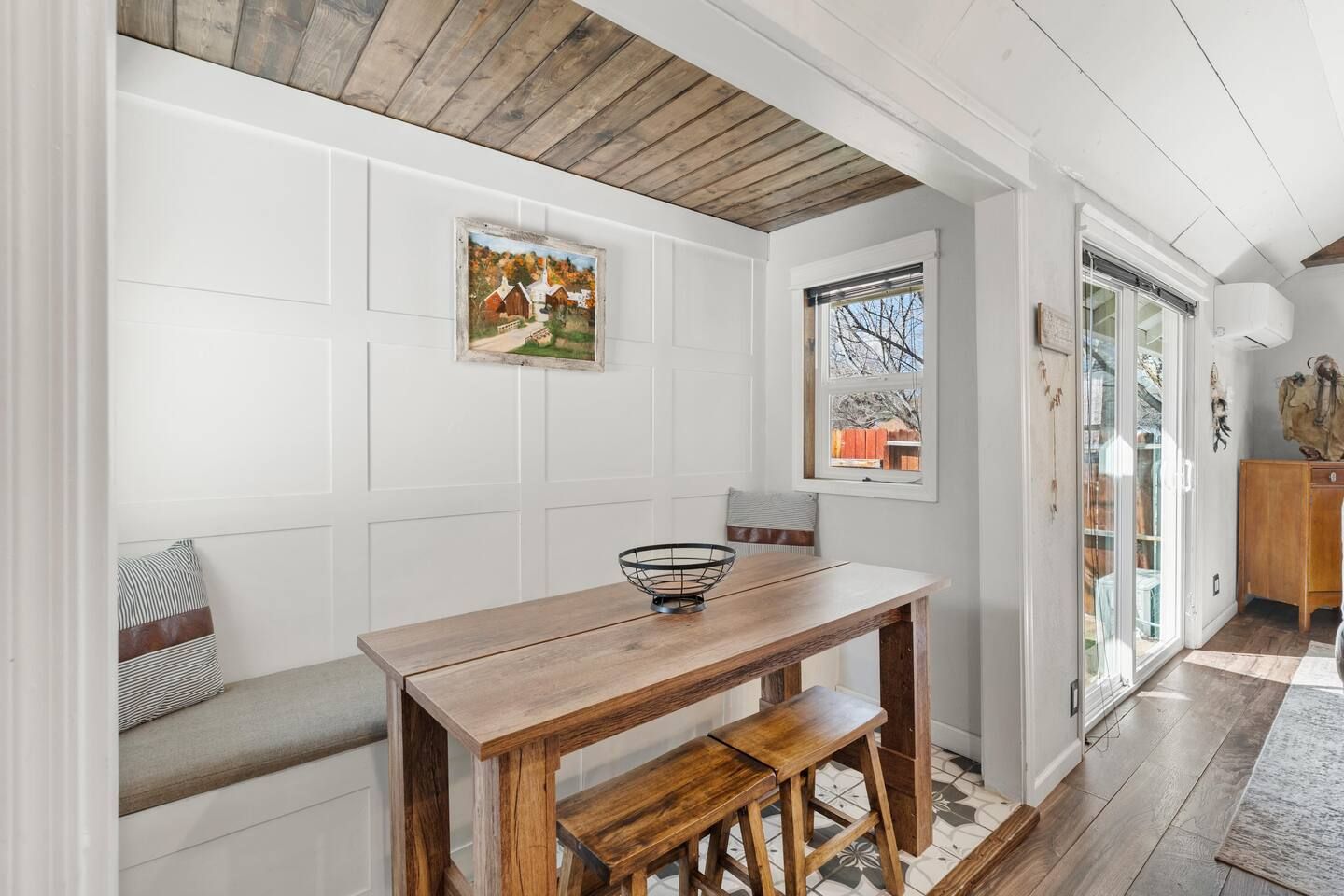 A dining area with a wooden table and bench, a framed picture on the wall, and a window with a view of the outside.