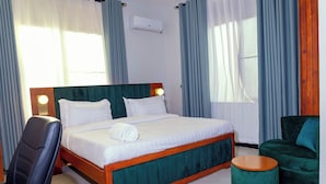 A hotel room with a bed, a chair, a round ottoman, and a wall-mounted air conditioner. - Amani Residence Entebbe (Entebbe)