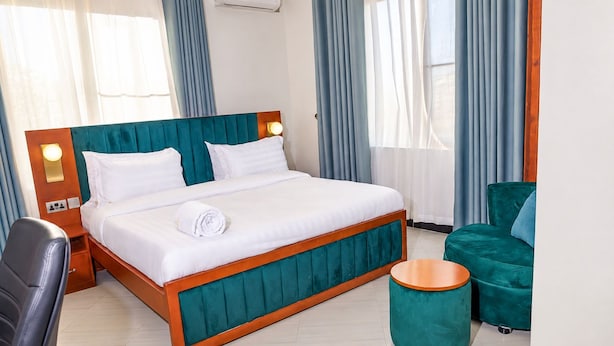 A hotel room with a bed, a chair, a round ottoman, and a desk with a lamp. - Amani Residence Entebbe (Entebbe)