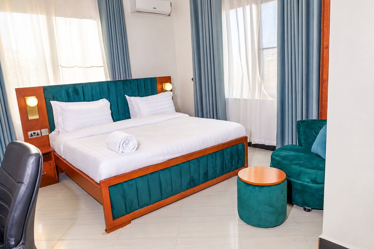 Ein Hotelzimmer mit einem Bett, einem Sessel, einem runden Hocker und einem Schreibtisch mit einer Lampe.