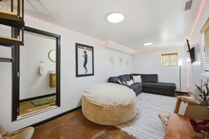 A modern living room with a dark grey sofa, a round ottoman, a large mirror, and a framed poster on the wall.