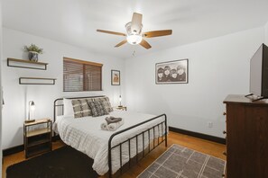 A bedroom with a bed, bedside tables, a ceiling fan, a window with blinds, a wall-mounted shelf with a plant, a framed picture, and a television on a wooden cabinet.