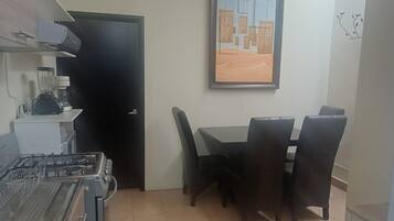 Cocina con estufa a gas, licuadora y microondas. Hay una mesa de comedor con cuatro sillas. Se ve una puerta a la izquierda, y un cuadro enmarcado cuelga de la pared sobre la mesa.
