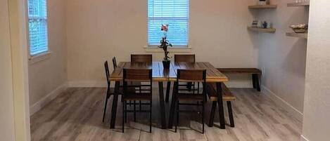 Un coin repas avec une table et des chaises en bois, un banc et une fenêtre avec des stores.
