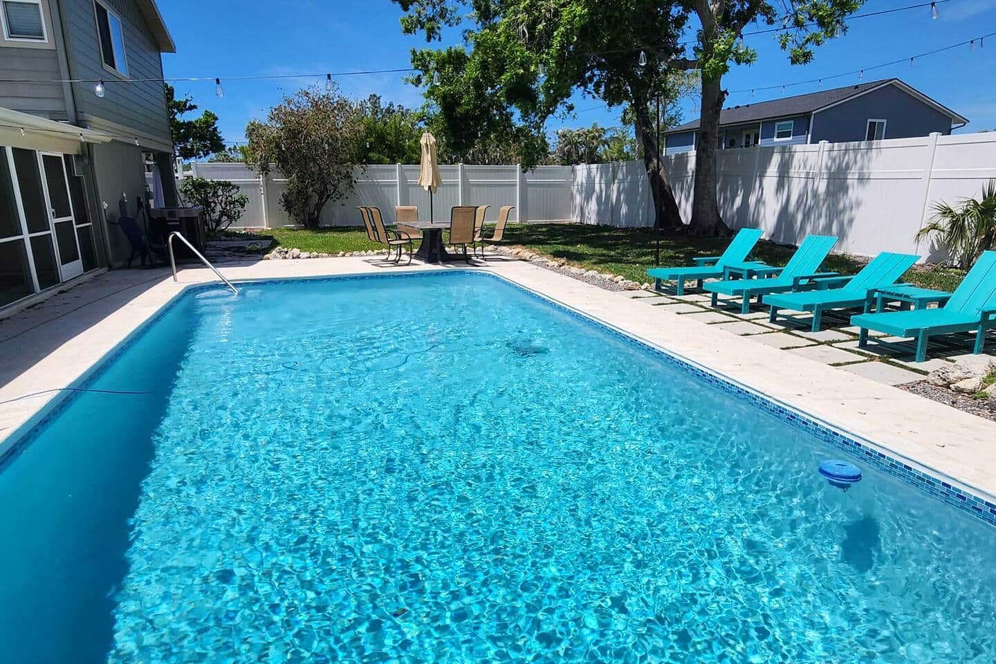 Ein rechteckiger Swimmingpool mit klarem, blauem Wasser, umgeben von einem weißen Zaun, mit Liegestühlen und Sonnenschirmen.