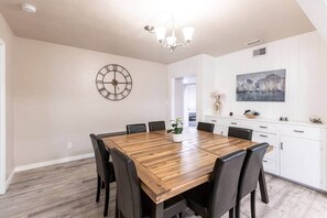 Uma área de refeições com mesa de madeira e cadeiras pretas, um relógio de parede e um quadro sobre um armário branco.