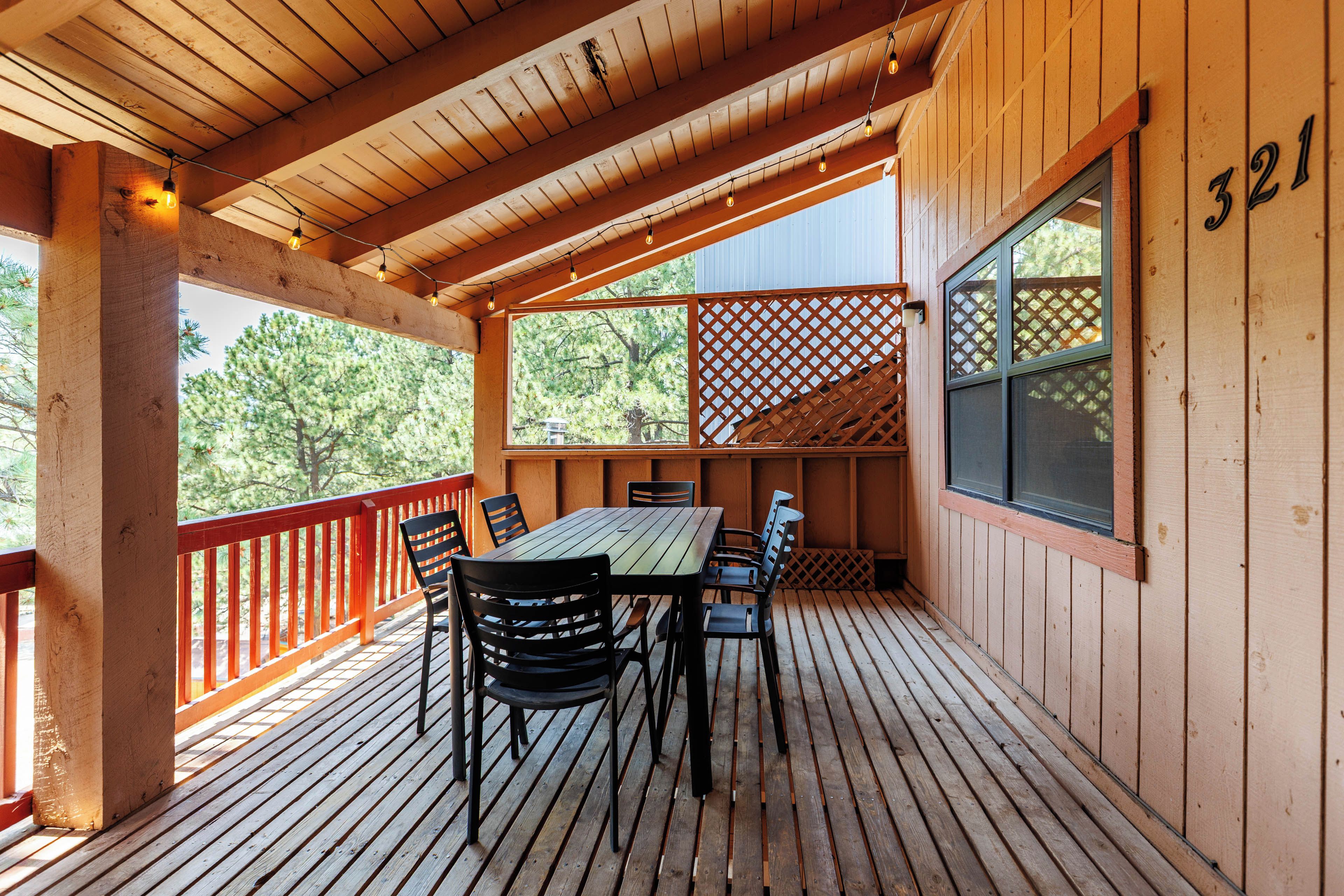 Una terraza de madera con mesa y sillas, barandilla roja y un cerramiento ajardinado.