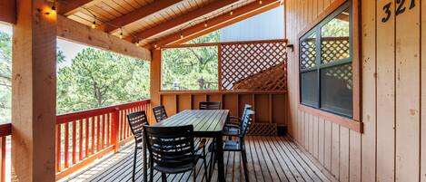 Una terraza de madera con mesa y sillas, barandilla roja y un cerramiento ajardinado.