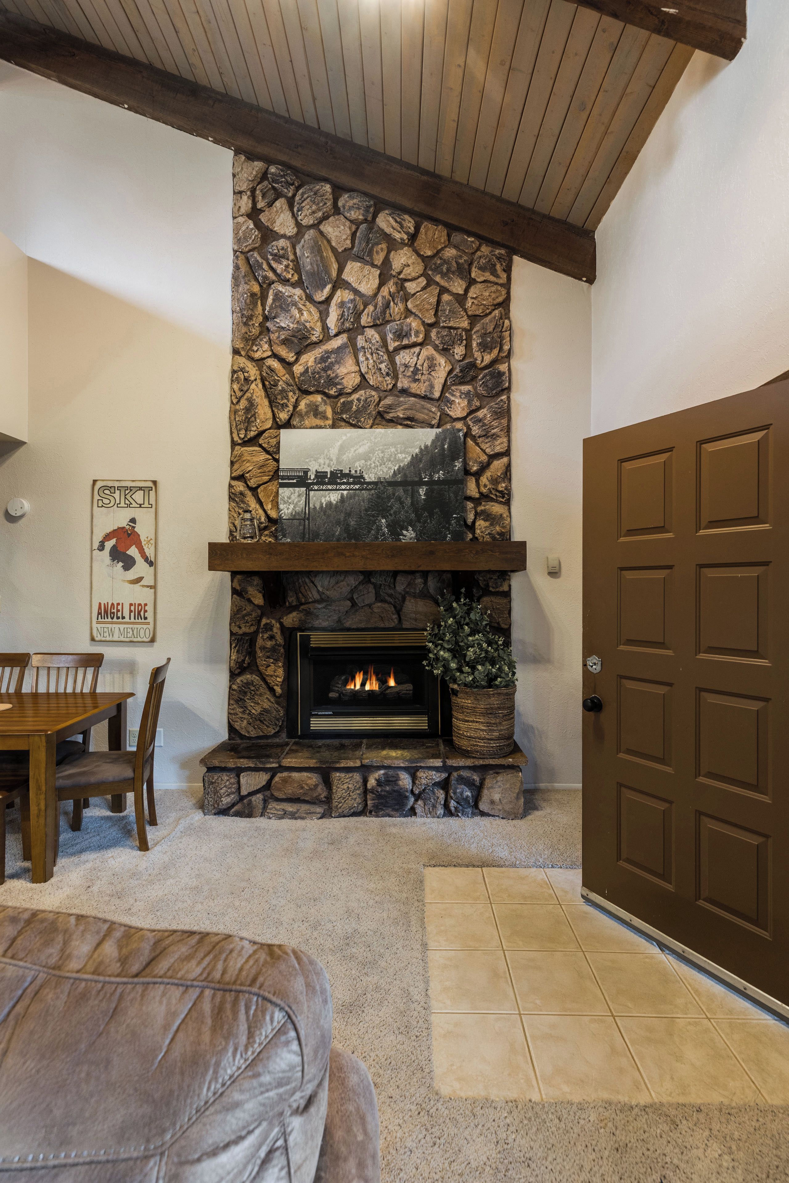 A stone fireplace with a framed picture above it, a dining table with chairs, and a brown door.