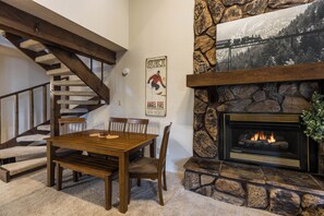A dining area with a wooden table and chairs, a fireplace with a stone surround, and a staircase with wooden beams. - Pine Top Loft - Walk to Lifts, Ski Locker, Deck (Angel Fire)