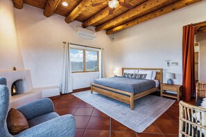 Quarto com teto de madeira, cama com cabeceira, mesa de cabeceira, cadeira, janela com cortinas e lareira.