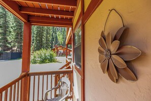 A wooden porch with a metal flower decoration on the wall, a chair, and a view of a forested area with a building in the background. - Tenderfoot Retreat - Creekside, Near Ski & Town (Red River)