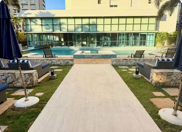 Un área de piscina con sillones reclinables y un sendero de piedra que conduce a un edificio con grandes ventanales.